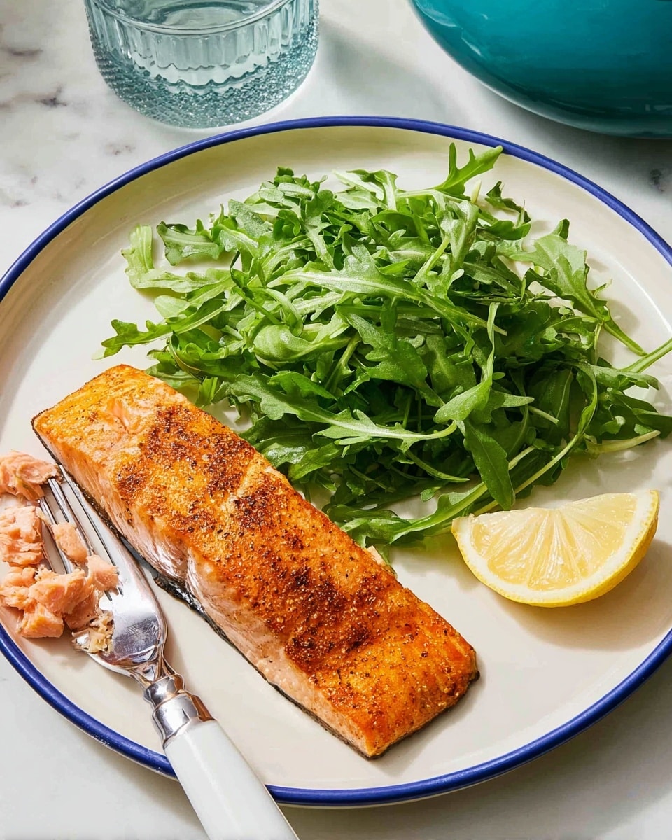 A white plate with a thin blue rim holds a cooked salmon fillet on the left side, showing a golden-orange color with a slightly crispy, seasoned top layer. Next to it, on the right side of the plate, is a mound of fresh, bright green arugula leaves, adding a leafy texture. Above the salmon piece, there is a bright yellow lemon wedge resting partly on the arugula. A silver fork with a white handle rests on the plate’s bottom left edge, with small chunks of salmon on its prongs. The plate sits on a white marbled surface, and a glass and a teal pot are faintly visible in the background. photo taken with an iphone --ar 4:5 --v 7