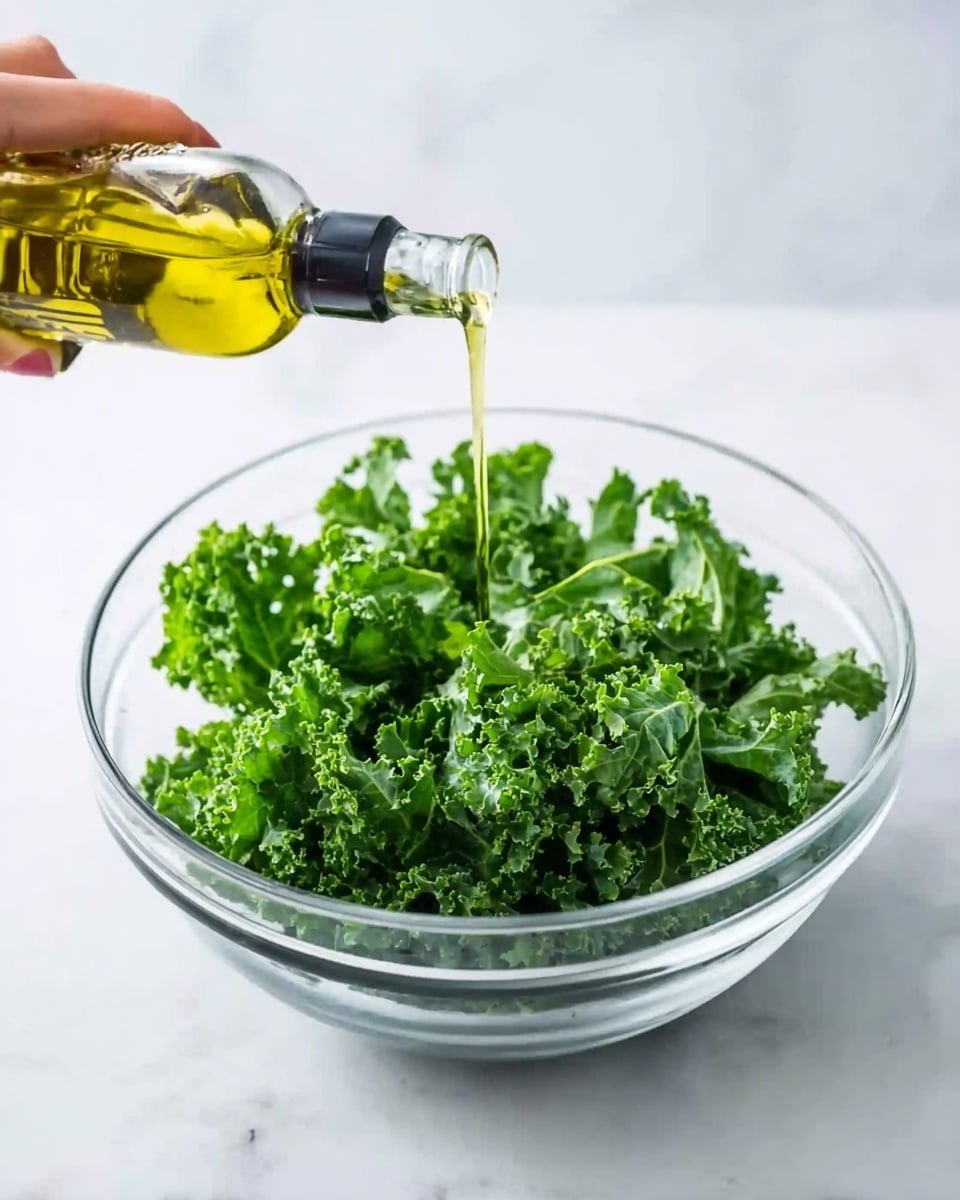 A clear glass bowl filled with fresh, bright green kale leaves that look crispy and slightly curly. A woman's hand is pouring golden olive oil from a glass bottle onto the kale. The bowl is placed on a white marbled surface that adds a clean and fresh look to the scene, photo taken with an iphone --ar 4:5 --v 7