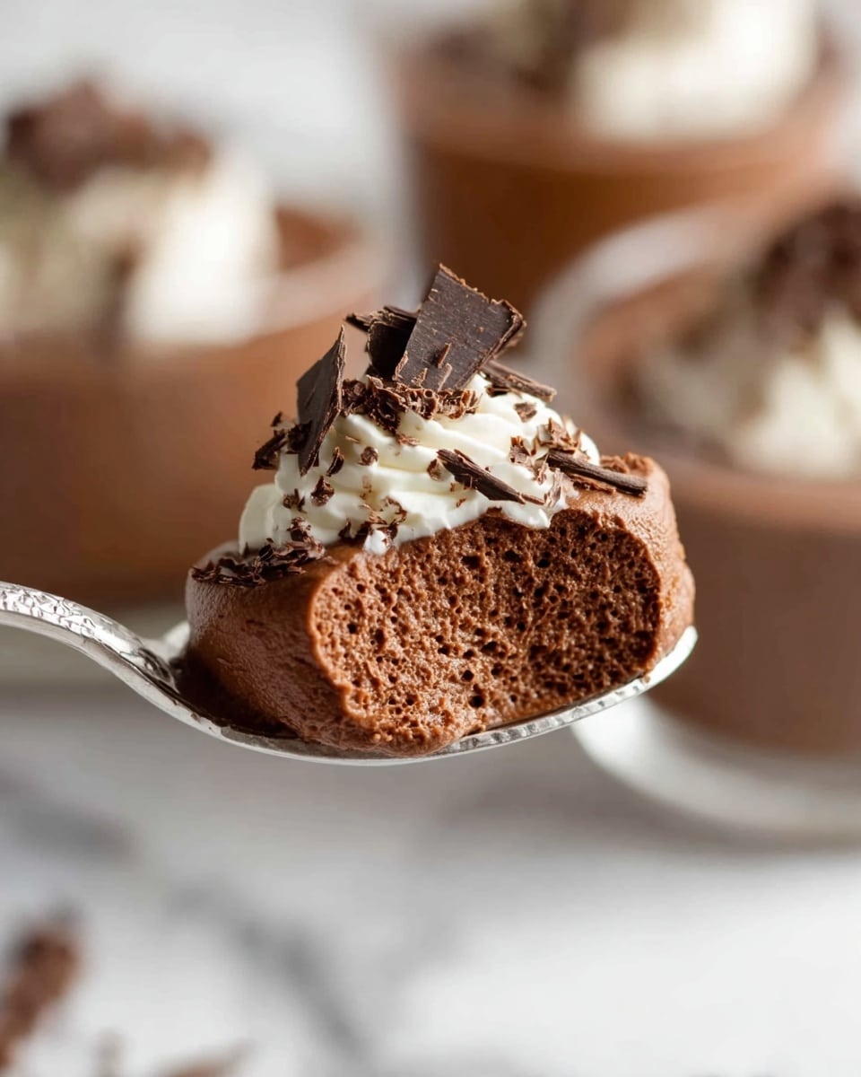 A close-up of a spoon holding a single scoop of light, airy chocolate mousse that has a smooth, fluffy texture with tiny air bubbles visible. The mousse is dark brown and topped with a small swirl of white cream and thin, dark chocolate shavings scattered on top. In the blurred background, there are white containers filled with more chocolate mousse topped with cream and chocolate shavings, all placed on a white marbled surface. photo taken with an iphone --ar 4:5 --v 7