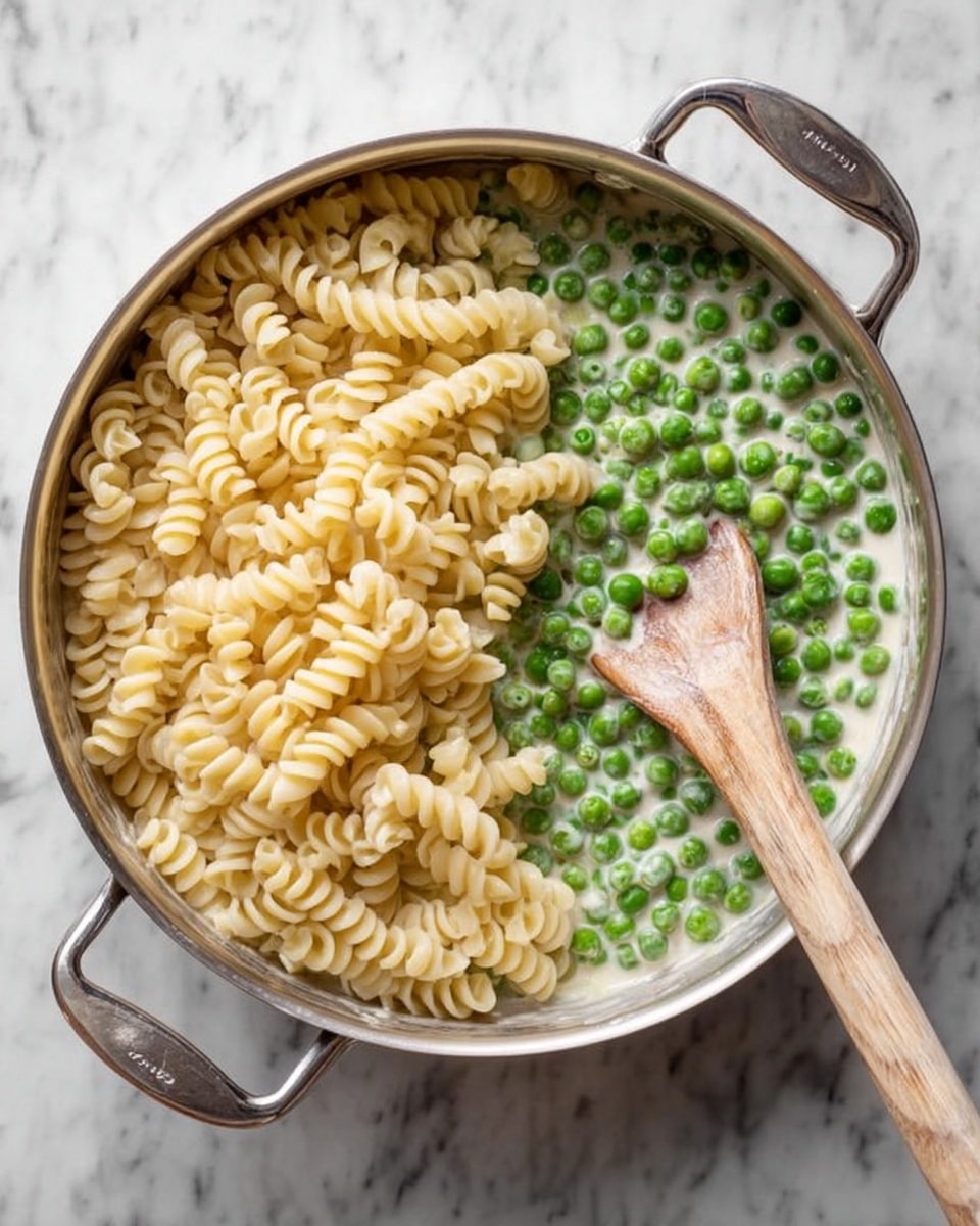 A silver pan with two handles is shown from above, filled halfway with small green peas in a white creamy sauce on the bottom half. Over the peas, there is a heap of pale yellow spiral pasta piled up on the left side. A long wooden spoon lies diagonally across the pan, resting on the peas and pasta, with a smooth texture. The pan is placed on a white marbled textured surface. photo taken with an iphone --ar 4:5 --v 7