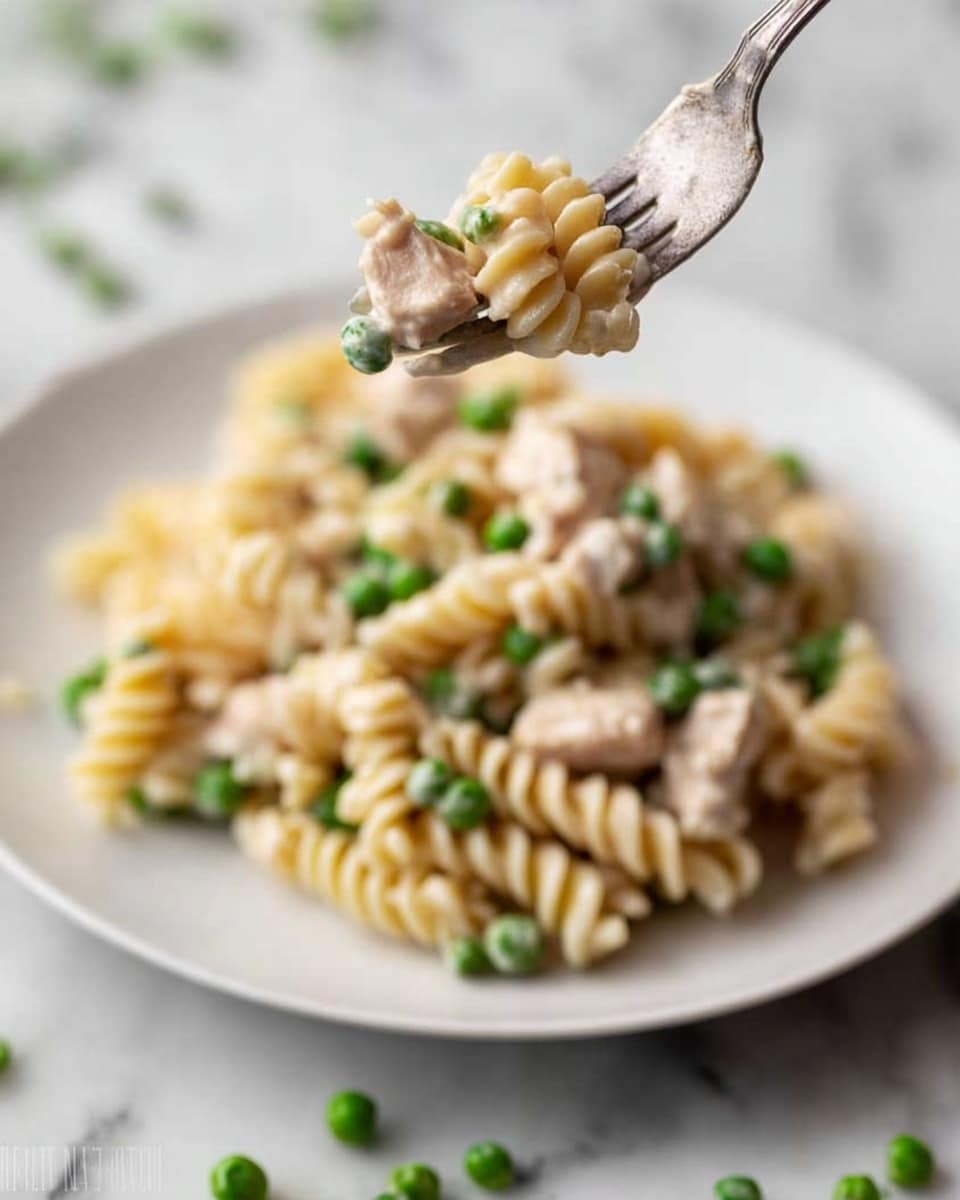 A close-up of a white plate filled with creamy pasta, showing one forkful lifted above the plate. The pasta is curly and coated in a light cream sauce, mixed with small green peas and tender pieces of cooked chicken scattered throughout. The sauce looks smooth and slightly shiny, covering the pale pasta and bits of chicken evenly. The background is a white marbled surface with some scattered peas and pasta around the plate. Photo taken with an iphone --ar 4:5 --v 7
