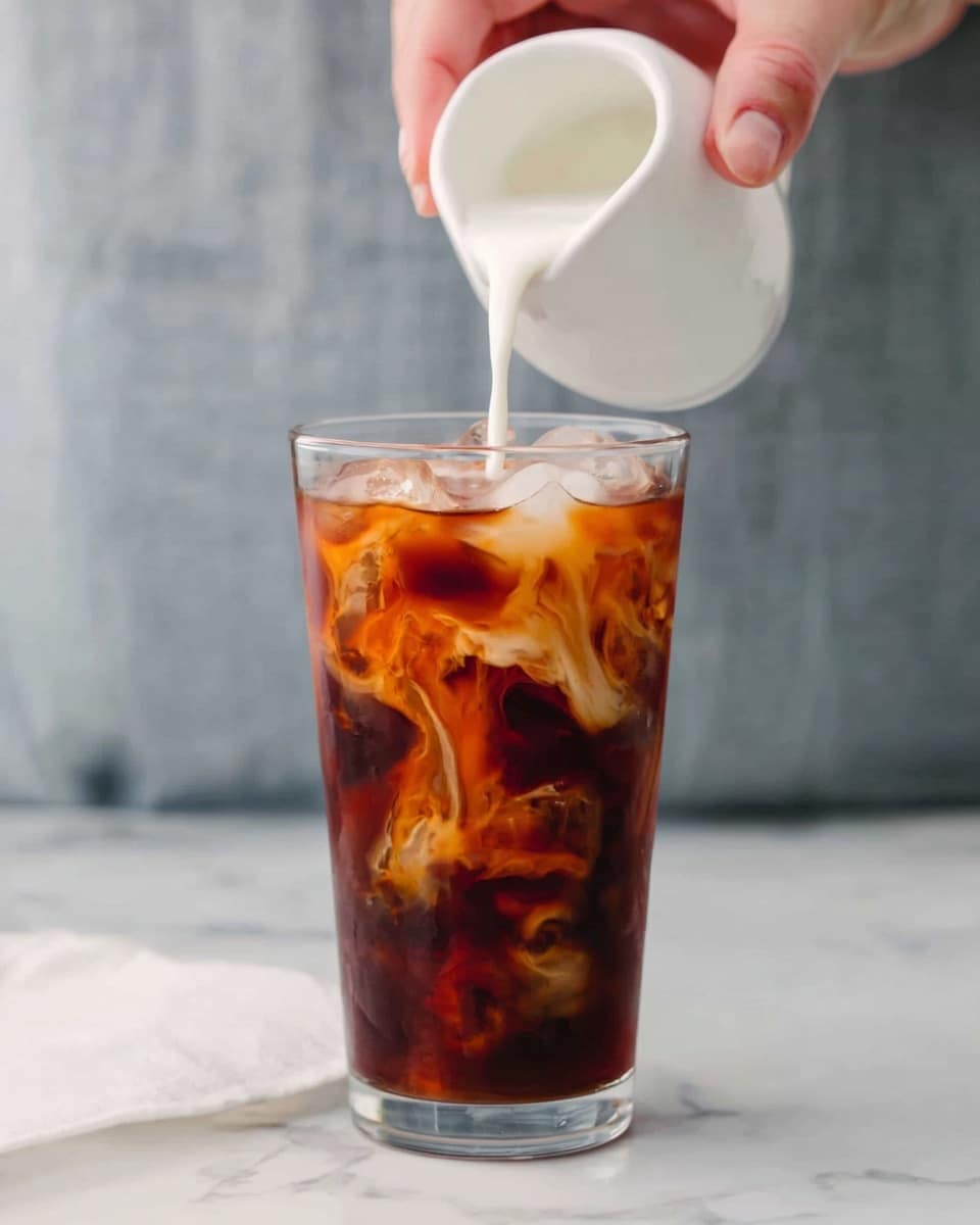 A tall clear glass filled with dark brown iced coffee and ice cubes inside. A woman's hand is pouring white milk from a small white pitcher into the glass, creating a swirling pattern as the milk mixes with the coffee. The background is softly blurred with a white marbled surface below the glass. photo taken with an iphone --ar 4:5 --v 7