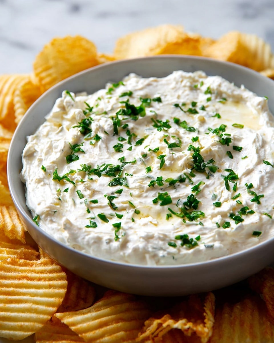 A close-up view shows a grey bowl filled with a creamy white dip with a thick and smooth texture. The dip has a swirled surface, topped with finely chopped green herbs, scattered evenly in the center and around the middle. The bowl sits on a white marbled surface, surrounded by ridged yellow potato chips, partially visible around the edges. The dip's richness contrasts with the chips' crisp texture, making a simple and inviting snack setup photo taken with an iphone --ar 4:5 --v 7