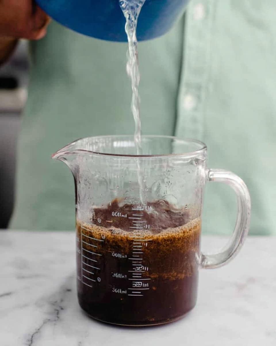 A clear glass measuring cup filled with dark brown coffee grounds at the bottom and water being poured from a blue container into the measuring cup, with the water creating a wet layer on top of the grounds. The cup is placed on a white marbled surface, and a person wearing a light green shirt is blurred in the background. The measuring cup shows volume markings on its side in black. photo taken with an iphone --ar 4:5 --v 7