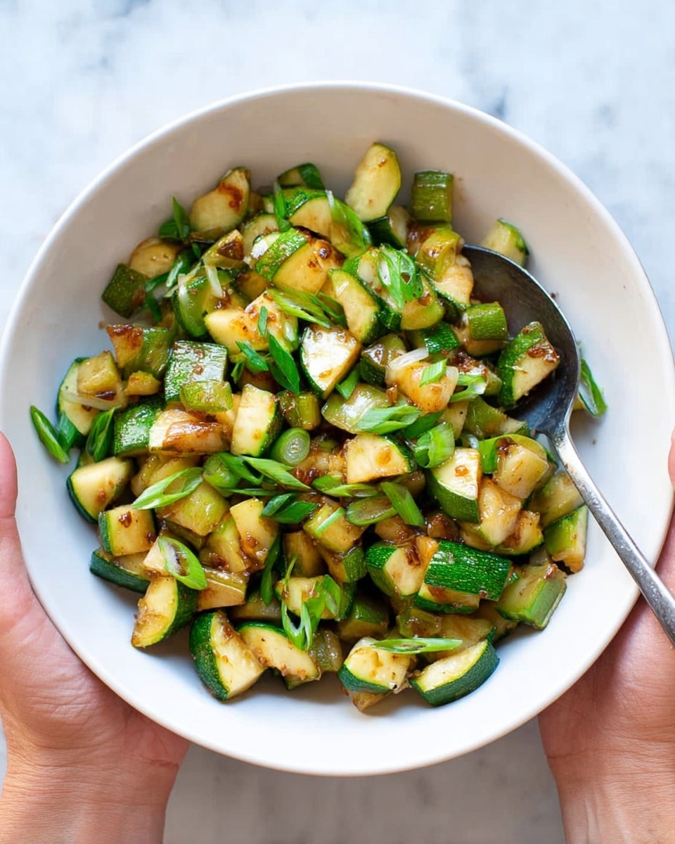 A white bowl is filled with cooked zucchini pieces. The zucchini is cut into small chunks and slices, with a mix of green skin and golden-brown cooked edges showing a slight crisp. Some green onion slices are scattered on top, adding a fresh green color. A silver spoon rests inside the bowl, and the bowl is held by two woman's hands over a white marbled surface. The light shows the shiny, soft texture of the cooked zucchini. photo taken with an iphone --ar 4:5 --v 7