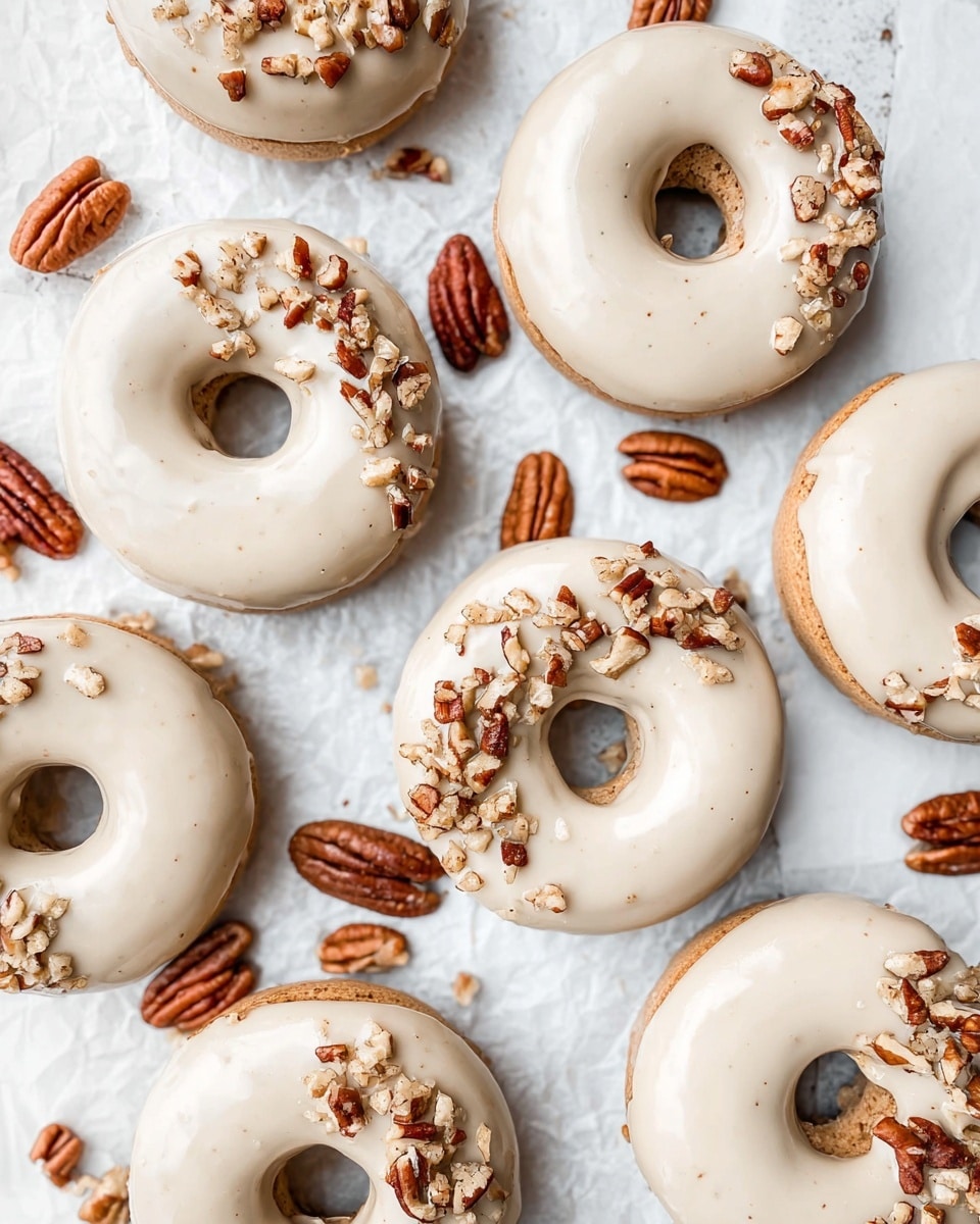 The image shows a group of round donuts arranged closely on a white marbled surface covered with crinkled white paper. Each donut has two layers: a base layer of light brown dough and a top layer of smooth, light beige glaze. Some of the donuts have small pieces of chopped pecans sprinkled on one side, adding a rough texture and dark brown color contrast to the creamy glaze. Whole and halved pecans are scattered randomly around the donuts on the paper. The overall feel is neat and inviting with a soft color palette. photo taken with an iphone --ar 4:5 --v 7