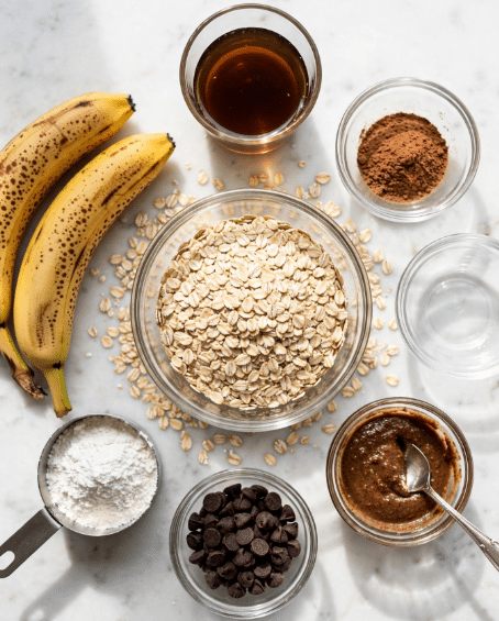 The image shows a clear glass bowl full of light brown oats in the center, surrounded by six small clear bowls and two brown ripe bananas with brown spots on the white marbled surface. Clockwise from the top are a glass cup with dark amber syrup, a small bowl with brown powder, an empty small bowl, a small bowl with dark brown almond butter and a silver spoon inside, a small bowl with dark brown chocolate chips, and a small metal measuring cup with white powder inside. Photo taken with an iphone --ar 4:5 --v 7