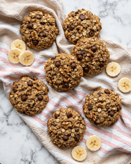 The image shows several round cookies made of oats and chocolate chips, scattered on a soft, beige towel with white and light pink stripes. The cookies are chunky and textured, with visible oats and dark brown chocolate chips spread throughout. Small pieces of walnut are also mixed in, adding to the texture. There are a few slices of banana placed around the cookies, which are pale yellow with a soft texture. The whole scene is set on a white marbled surface. photo taken with an iphone --ar 4:5 --v 7