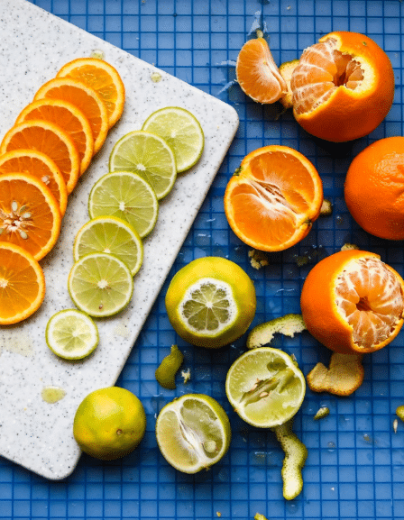 The image shows a group of peeled citrus fruits with their flesh exposed, arranged on a blue grid surface. There are primarily two types of fruits visible: bright orange ones with orange flesh and yellow-greenish ones with light green flesh. Each fruit has a patch where the peel is removed to reveal the juicy sections inside. On the left side, there is a white speckled cutting board with thin, round slices of these citrus fruits neatly placed on it. The fruits are scattered with some cut in half and some showing patterns in their peeled areas. The overall look is fresh and colorful, set on a clean blue grid background. photo taken with an iphone --ar 4:5 --v 7