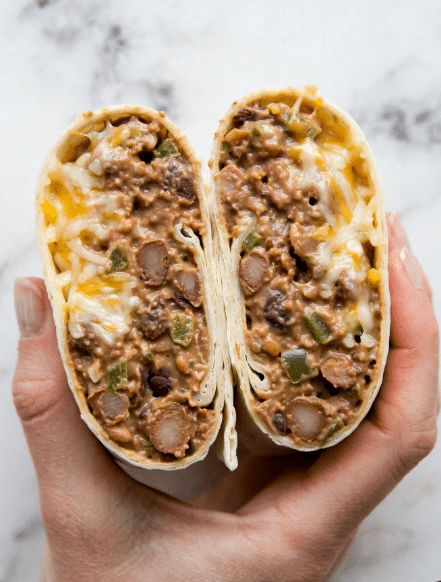 A close-up view of two halves of a wrap held by a woman's hand, showing three clear layers inside each half. The outer layer is a soft white tortilla that wraps the filling. The middle layer is thick, brown, and creamy with visible chunks of beans and small pieces of green peppers. The top layer has melted shredded cheese in white and yellow shades mixed into the brown filling, adding a smooth and cheesy texture. The background is a white marbled surface. Photo taken with an iphone --ar 4:5 --v 7
