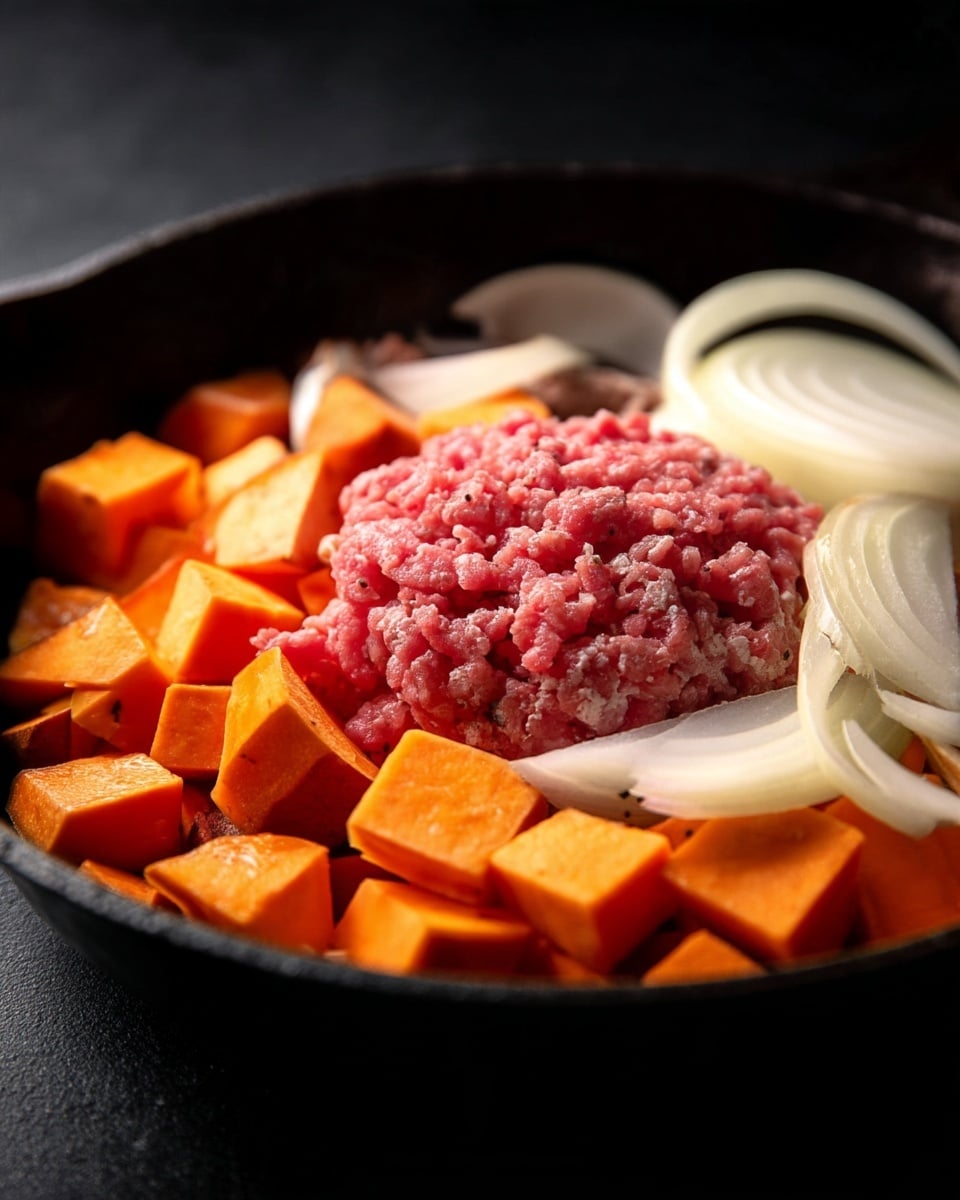 Inside a dark cast iron pan, there are three main layers of ingredients. The bottom layer is made of small orange sweet potato cubes that have a rough, matte texture. On top of that, slices of white onions with a smooth and slightly shiny surface are spread unevenly, their layers visible and slightly curved. The top layer features raw, pink ground meat placed mostly in the center, with its coarse texture contrasting with the vegetables around it. The pan sits on a dark surface. photo taken with an iphone --ar 4:5 --v 7