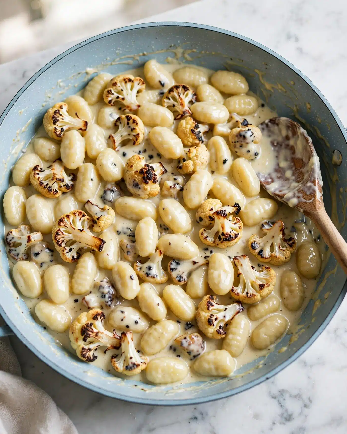 A large light blue pan filled with about two layers of creamy gnocchi mixed with pieces of browned cauliflower. The gnocchi are pale yellow with a smooth texture, while the cauliflower is slightly charred with brown edges, adding specks of darker color throughout. A wooden spoon is partially resting on the right side of the pan, coated with the creamy sauce that glistens slightly. The pan sits on a white marbled surface. photo taken with an iphone --ar 4:5 --v 7