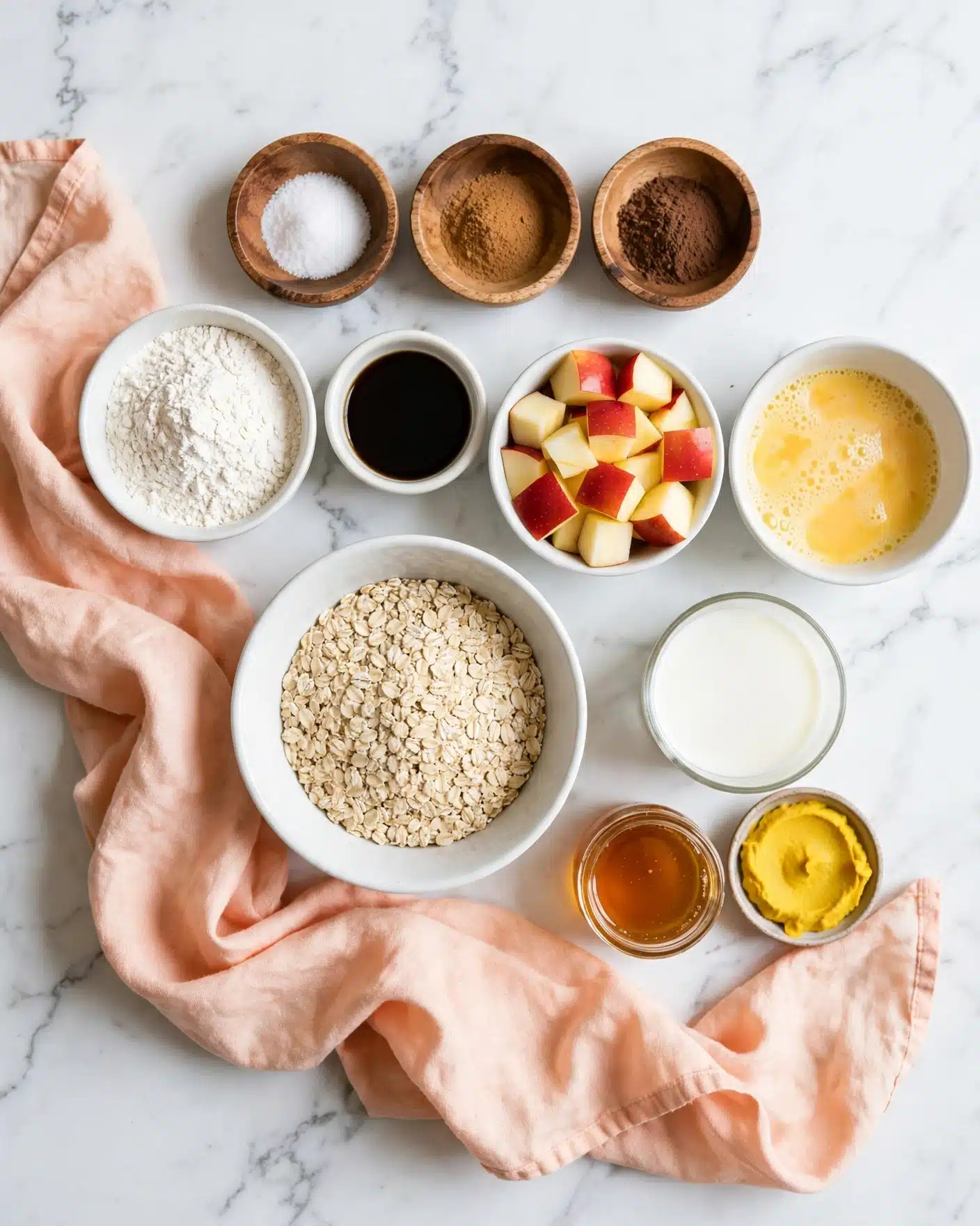 The image shows several small white bowls and wooden containers arranged neatly on a white marbled surface. The center features a white bowl filled with dry rolled oats, light beige in color with a rough texture. To the top right of the oats is a white bowl filled with small cubes of red and yellow apple, colorful and fresh-looking. Near the apple, there is a small white bowl with yellow beaten eggs, slightly foamy on top. Above the oats are three small wooden bowls containing salt and two powdered spices of light brown and dark brown colors. To the top left side of the oats is a small white bowl of white flour, a small white bowl with a dark liquid, and a glass cup filled with white milk. Near the glass are smaller containers with a honey-like amber liquid and a bright yellow paste. A soft peach-colored cloth is gently spread around the bowls, adding softness to the setup. Photo taken with an iphone --ar 4:5 --v 7