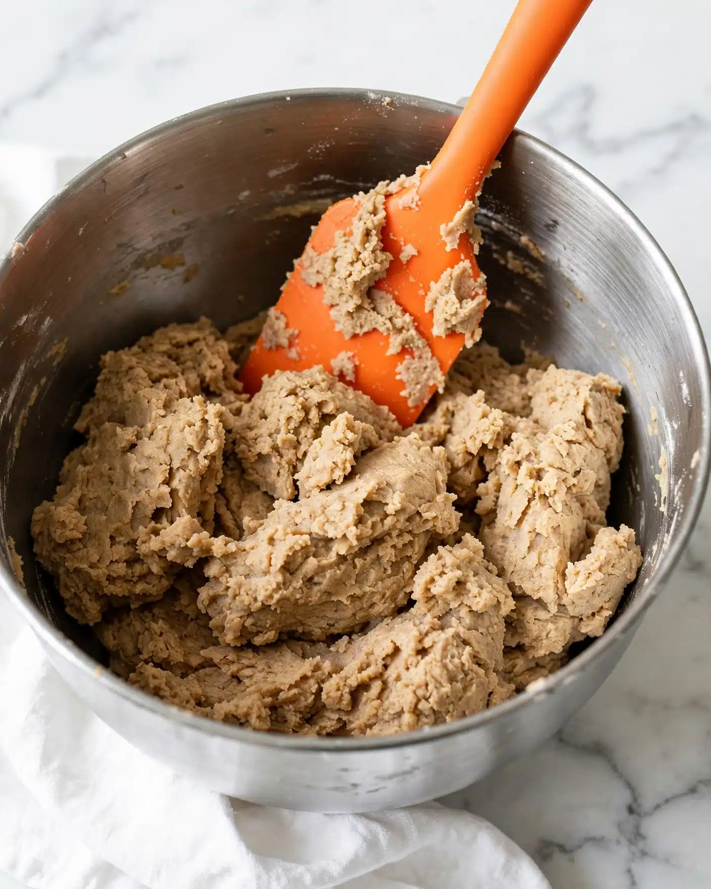 A silver mixing bowl filled with light brown dough that looks rough and thick, sitting on a white marbled surface. An orange silicone spatula is inside the bowl, resting at an angle with some dough stuck to it. A white cloth is partially visible below the bowl. The whole scene looks clean and bright with soft light. photo taken with an iphone --ar 4:5 --v 7