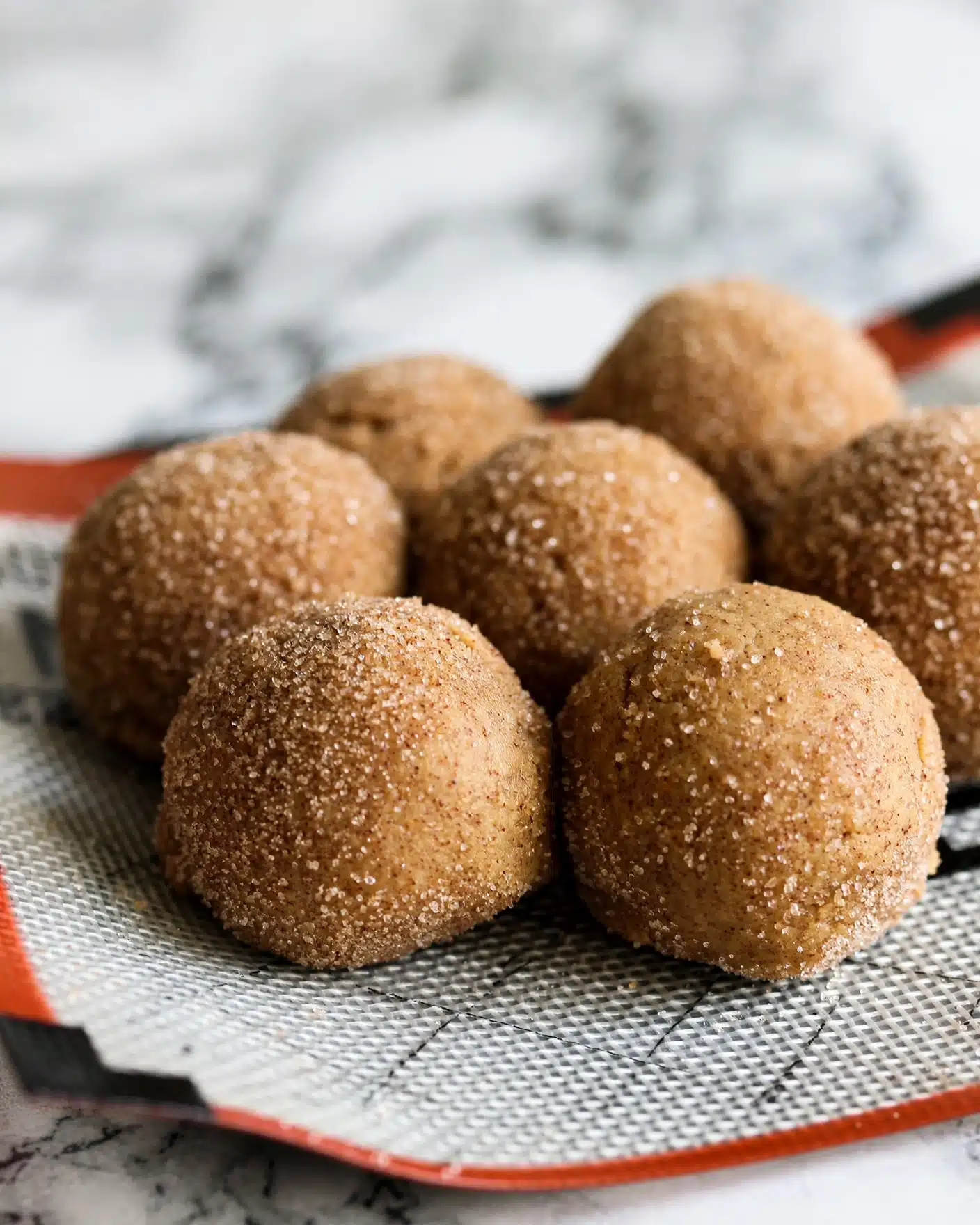 Pumpkin Snickerdoodles Recipe 4 The image shows a close-up of several round dough balls covered in a rough layer of granulated sugar mixed with cinnamon, giving them a sandy brown color with specks of darker cinnamon. They are all resting on a textured silicone baking mat that has a white base with a grid pattern and some red and black areas visible near the edges. The dough balls have a smooth, soft surface with a slight sparkle from the sugar coating. The background is a white marbled surface that contrasts softly with the warm tones of the dough balls. photo taken with an iphone --ar 4:5 --v 7