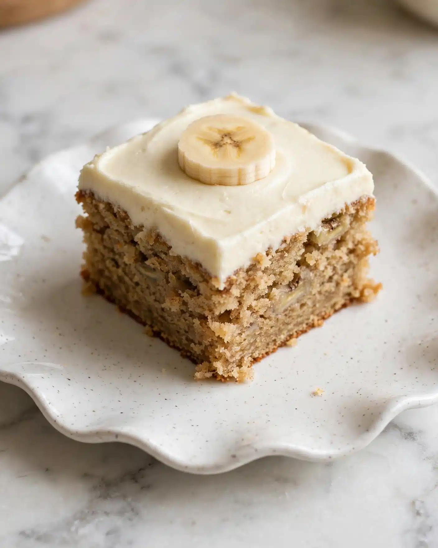 A single square piece of moist banana cake is shown on a white, slightly speckled plate with a wavy edge. The cake has one thick layer of light brown, soft, and crumbly texture with visible small banana bits. On top, there is one thick layer of smooth, creamy, off-white frosting. A round slice of banana lies flat on the frosting as decoration. The scene is set on a white marbled surface with soft natural light, giving a cozy and warm feeling. Photo taken with an iphone --ar 4:5 --v 7