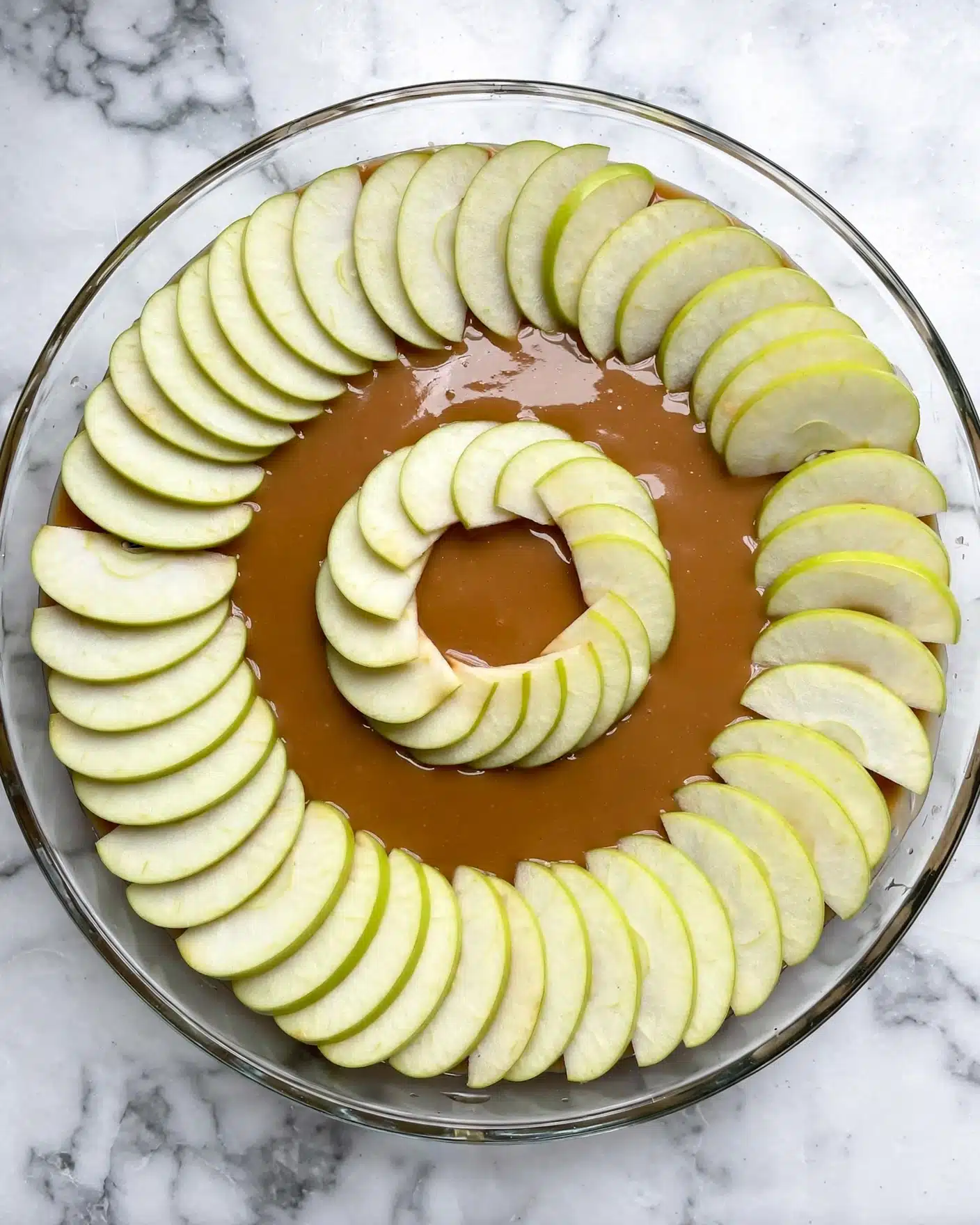 Caramel Apple Upside Down Cake Recipe 4 A round clear glass baking dish contains thin slices of pale green apples arranged in a circular pattern, with two rings of apple slices; one larger outer ring and a smaller inner ring, all placed on a glossy light brown sauce covering the bottom of the dish. The dish is set on a white marbled surface. photo taken with an iphone --ar 4:5 --v 7