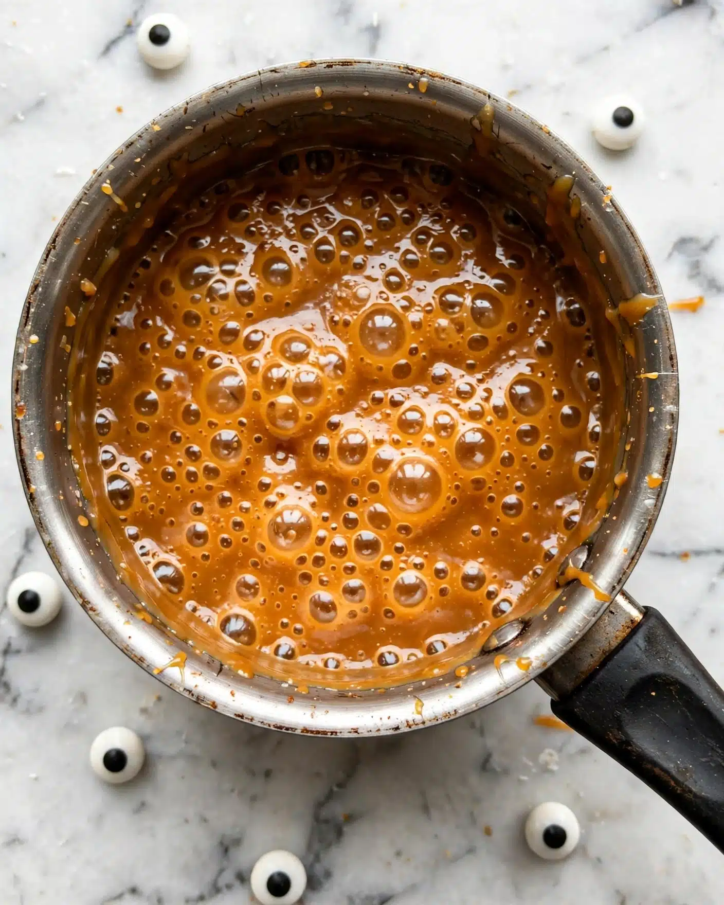 A close-up view of a metal pot filled halfway with thick, bubbling caramel sauce that is a rich amber color with shiny bubbles of different sizes covering the surface, resting on a white marbled countertop. Around the pot, there are several candy eyeballs scattered, some whole white with black centers, and others split open with a red splash pattern resembling fake blood. The pot has a dark handle attached and looks used, reflecting light gently. photo taken with an iphone --ar 4:5 --v 7