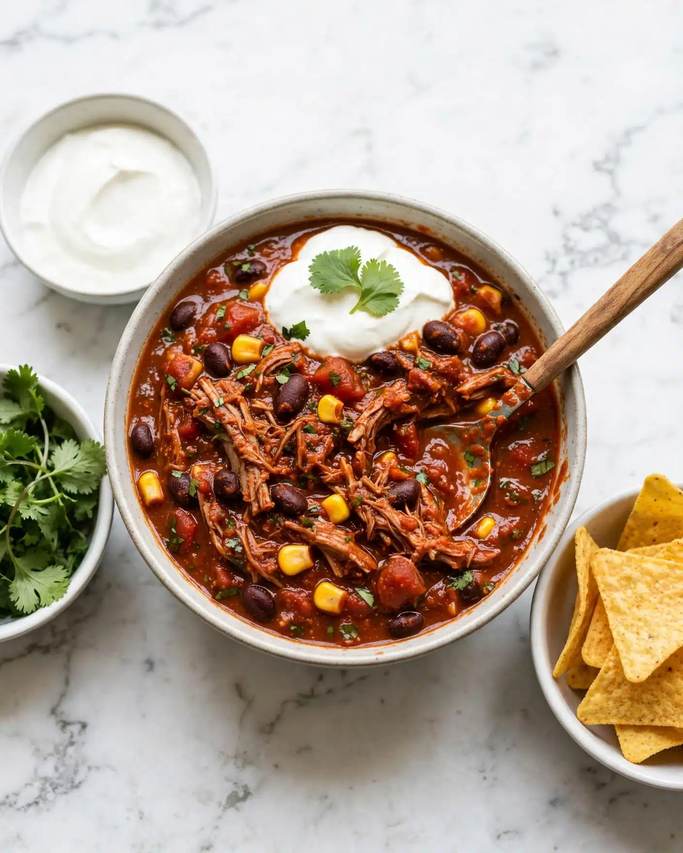 A bowl filled with thick tomato-based chili shows chunks of shredded meat, black beans, yellow corn, and diced tomatoes. The chili has green herb pieces scattered throughout and is topped with a dollop of white sour cream and a small green leaf. A spoon with a wooden handle rests inside the bowl, partially submerged in the chili. Around the bowl, there are three small white bowls, one filled with green leafy herbs, another with white sour cream, and the last one off to the side with yellow tortilla chips. All items sit on a white marbled surface. Photo taken with an iphone --ar 4:5 --v 7