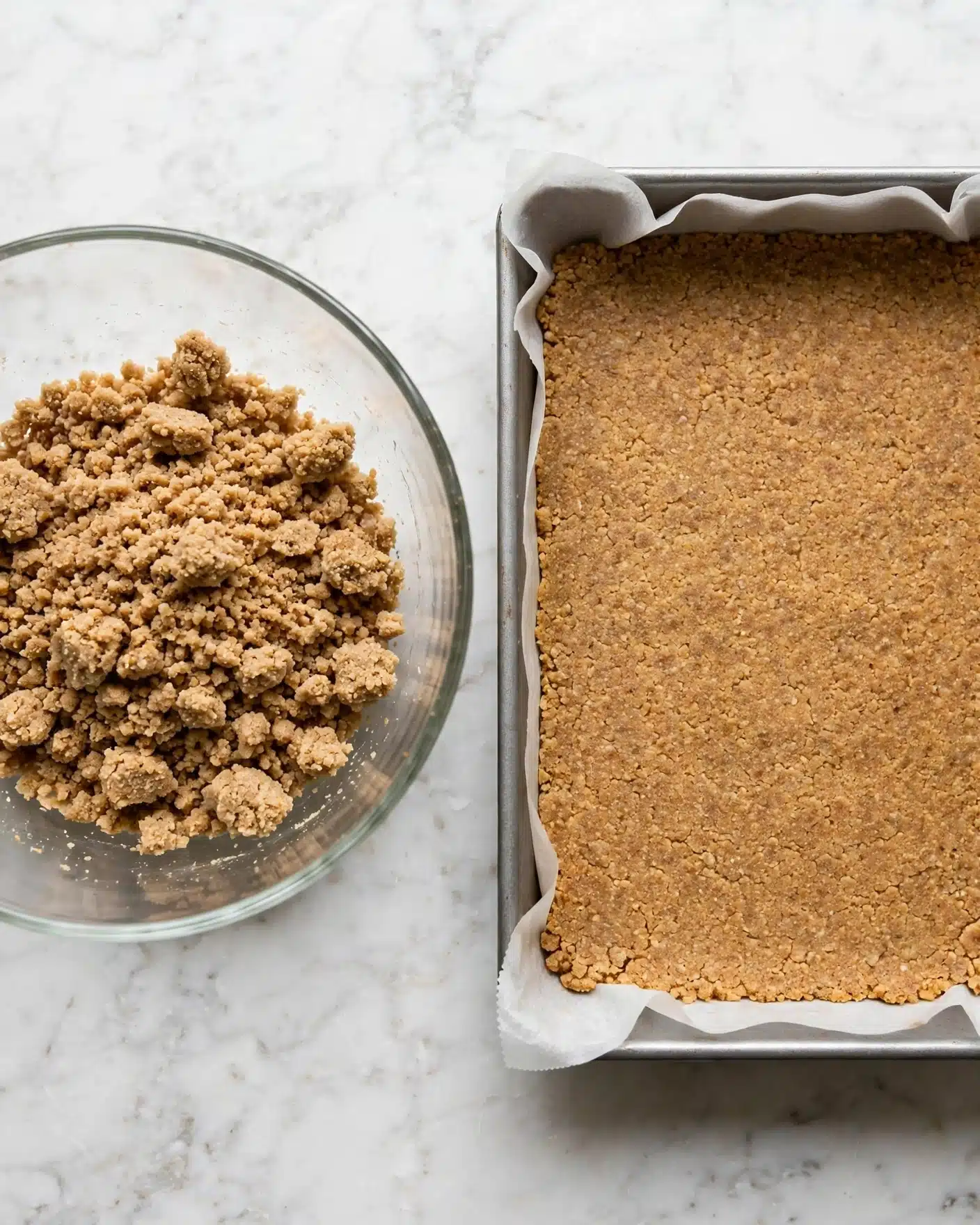 Pumpkin Caramel Cheesecake Bars with a Streusel Topping Recipe 4 The image shows two side-by-side photos on a white marbled surface. On the left, a clear glass bowl holds a crumbly mixture of light brown crumbs with a rough texture, filling the bowl unevenly. On the right, a baking pan lined with white parchment paper contains the crumb mixture pressed flat and even, spreading to cover the entire base of the pan in a single, compact layer of golden brown color and slightly grainy texture. photo taken with an iphone --ar 4:5 --v 7