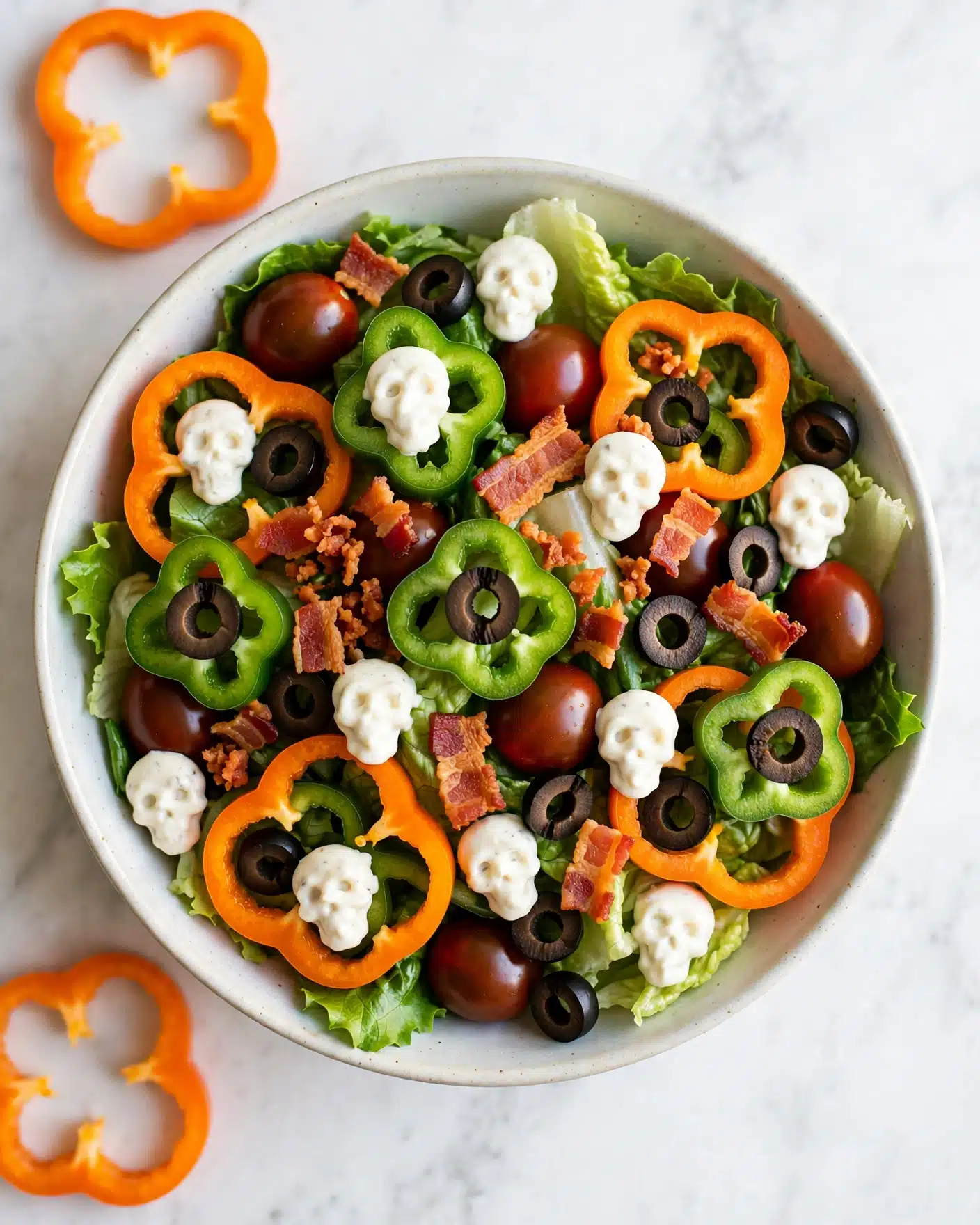 The dish is a colorful salad layered with leafy green lettuce at the bottom, topped with dark red cherry tomatoes, sliced black olives, and thin orange bell pepper rings. There are orange-red crispy bacon pieces scattered across, along with bright green bell pepper slices that have halves of black olives on top. Small dollops of white creamy dressing shaped like tiny skulls are placed evenly across the salad, creating a spooky effect. The salad sits on a white marbled surface. Photo taken with an iphone --ar 4:5 --v 7