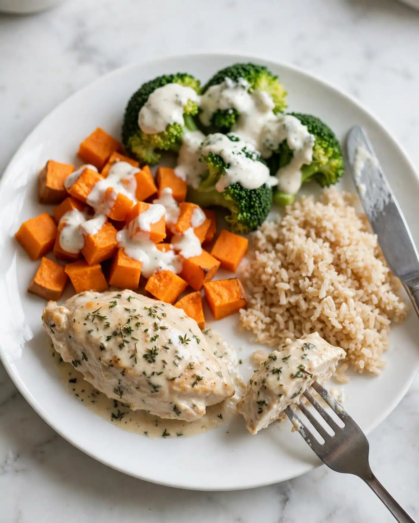 Smoked Salmon Dip with Everything Bagel Chips Recipe 5 The image shows a white plate with a meal divided into three sections: a creamy light beige chicken piece with green herb specks at the bottom left, bright orange sweet potato cubes topped with white sauce in the middle, and green broccoli florets also covered in white sauce at the top right. To the right of these ingredients is a serving of light brown rice with a slightly fluffy texture. A metal fork at the bottom right holds a piece of the creamy chicken, and a silver knife coated with some white sauce rests on the plate’s edge near the rice. The plate is set on a white marbled surface. Photo taken with an iphone --ar 4:5 --v 7