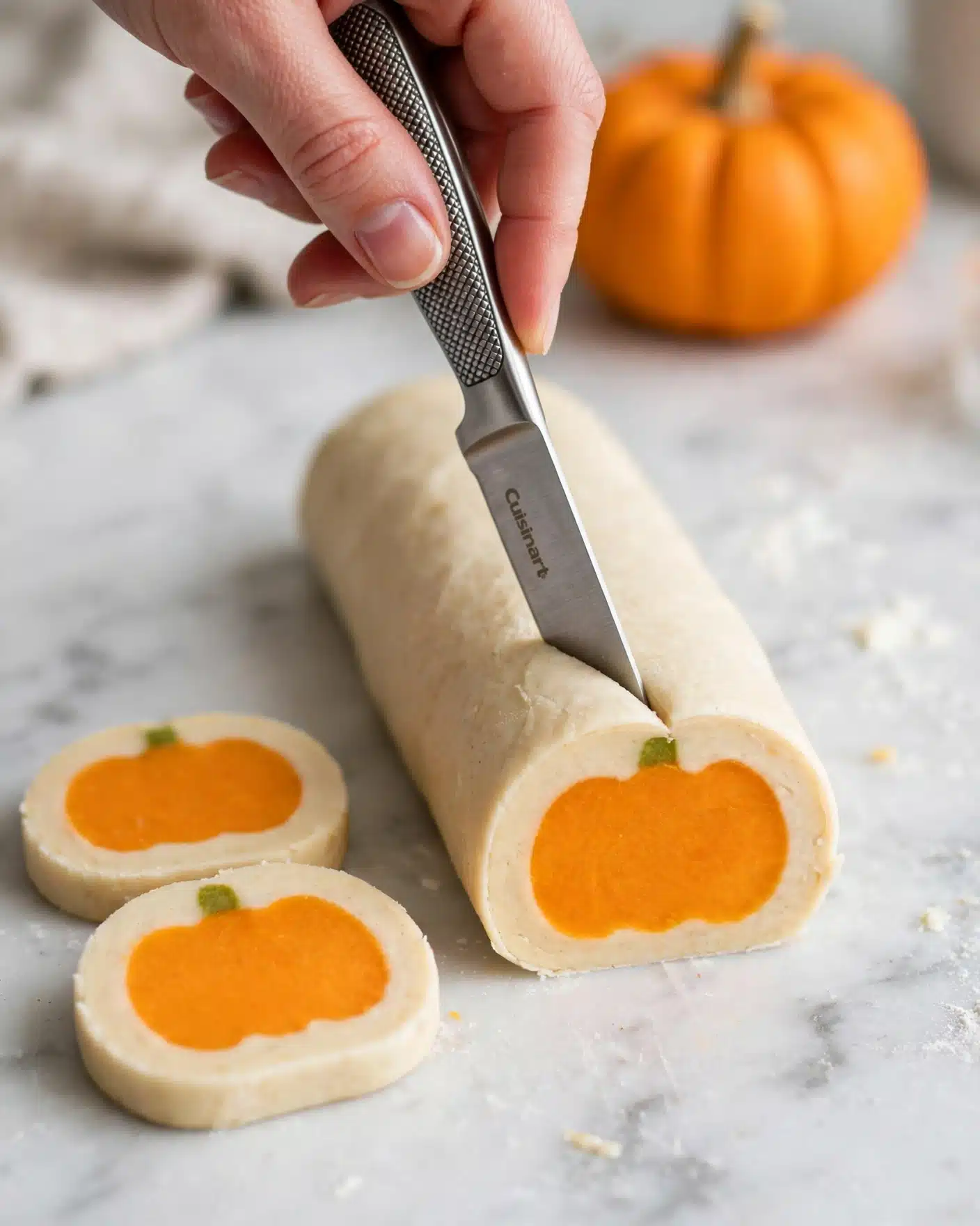 A white plate holds a neat stack of round sugar cookies, each having two layers: a soft creamy-white outer layer and a smaller, bright orange circle in the center representing a pumpkin shape. On top of the orange circle, there is a small green dot as the pumpkin stem. The cookies have a smooth surface but slightly grainy texture, and they are arranged overlapping with a focus on the front cookie. The plate is set on a black and white striped cloth with a blurred pumpkin in the background, all placed on a white marbled surface. photo taken with an iphone --ar 4:5 --v 7
