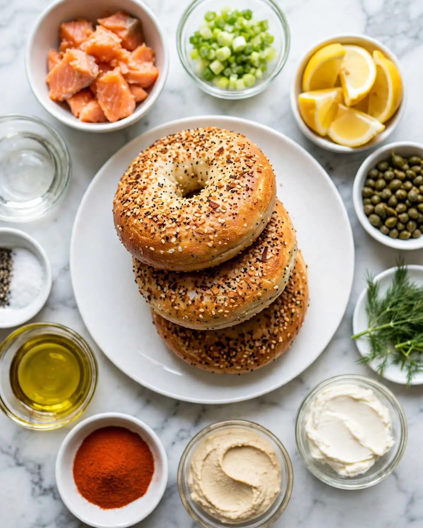 Smoked Salmon Dip with Everything Bagel Chips Recipe 4 A white plate holds three everything bagels stacked slightly on each other, showing their golden brown color with mixed sesame seeds and seasonings on top. Surrounding the plate on a white marbled surface are small white bowls and glass containers with colorful ingredients: pieces of cooked orange salmon in one bowl, light green chopped scallions in another, bright yellow lemon wedges in a white bowl, green capers in a white bowl, and fresh green dill on a small white dish. There are also glass bowls with creamy white spreads, one smooth light beige spread, a bowl of red-orange powder, a clear golden liquid, black pepper, white salt, and a clear liquid, all spread neatly around the bagels. Photo taken with an iphone --ar 4:5 --v 7