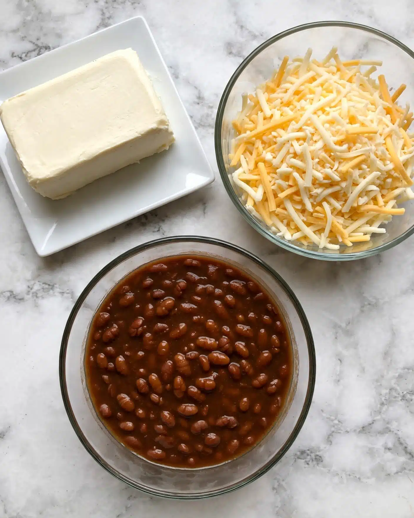 The image shows three items on a white marbled surface: at the top left, a rectangular block of cream-colored cream cheese on a white square plate; at the top right, a clear glass bowl filled with a mix of shredded light yellow and white cheese; at the bottom center, a clear glass bowl with dark brown baked beans in sauce, showing a thick texture and small beans throughout. Photo taken with an iphone --ar 4:5 --v 7