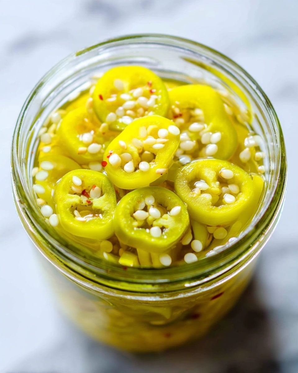The image shows a clear glass jar filled with sliced yellow-green pickled chili peppers. The peppers are thinly sliced into circular rings, with many white seeds scattered throughout the jar, visible from the top view. The jar is placed on a white marbled surface, and a white jar lid with a red inner ring is seen slightly out of focus in the background. The chili slices have a shiny, wet texture from the pickling liquid inside the jar. Photo taken with an iphone --ar 4:5 --v 7