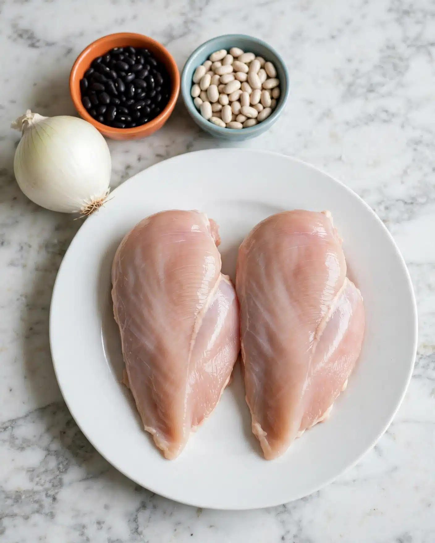 Two raw chicken pieces lie side by side on a white plate in the center of the image. To the top left of the plate, there is a white onion resting on a white marbled surface. Behind the onion, two small bowls sit side by side; the left bowl is orange and filled with black beans, while the right bowl is light blue and filled with white beans. The entire scene is set on a white marbled surface. Photo taken with an iphone --ar 4:5 --v 7