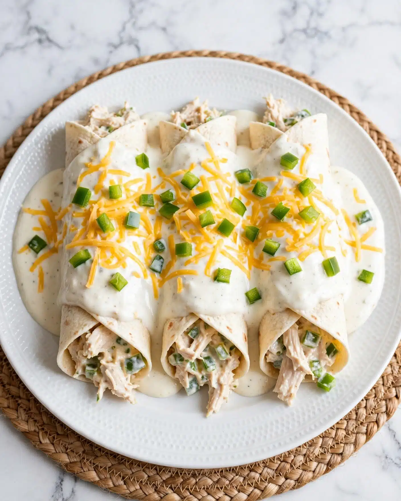 Creamy Chicken Enchiladas Recipe 5 The image shows three rolled tortillas filled with a light-colored chicken mixture that includes small green pepper pieces, all placed on a white plate with a subtle textured rim. The tortillas are topped with a thick white sauce that covers most of the dish, sprinkled with shredded yellow cheese and diced green peppers. The plate rests on a light brown woven mat, but the background is changed to white marbled texture. The overall look is creamy with pops of green and yellow from the peppers and cheese. photo taken with an iphone --ar 4:5 --v 7